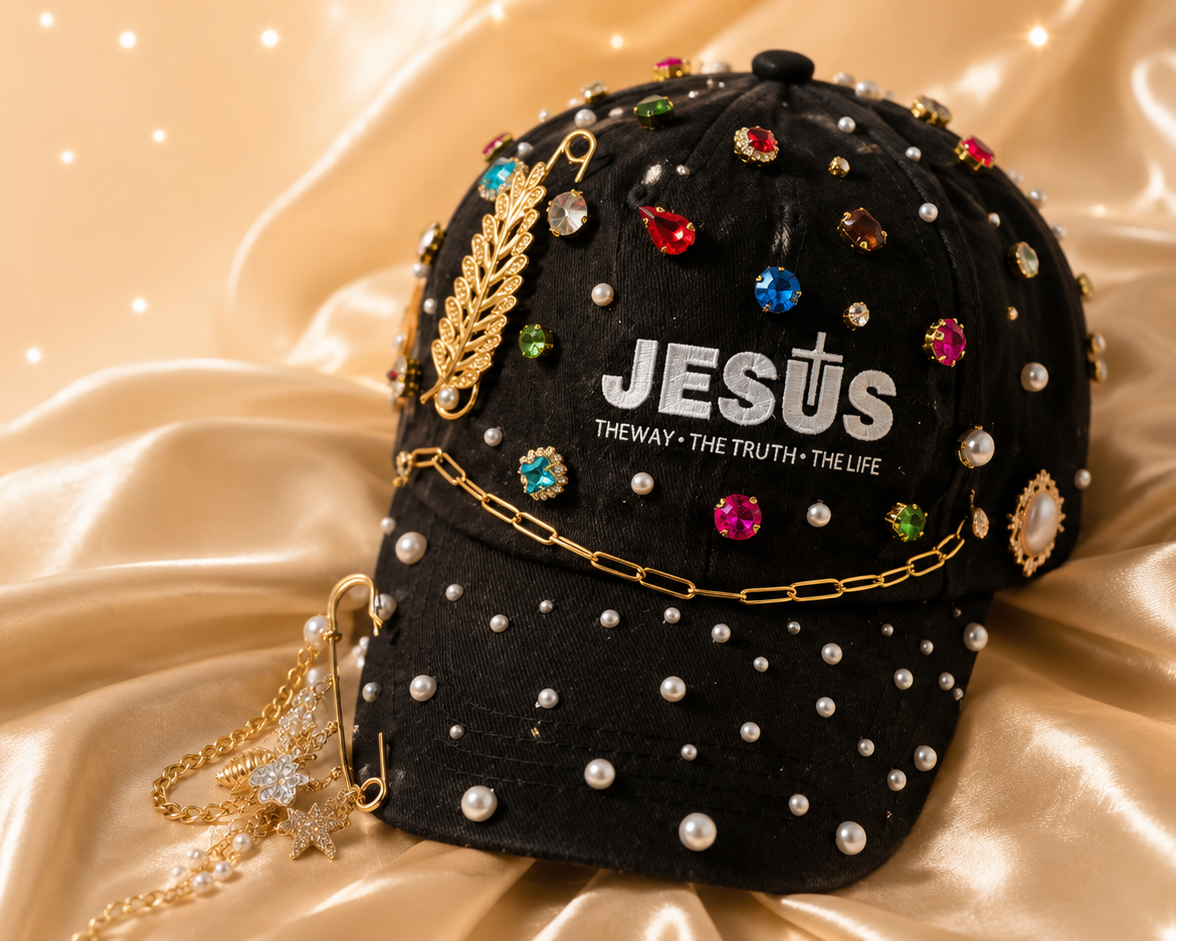 A black baseball cap embellished with white faux pearls, gold chains, and colorful rhinestones, featuring the text 'JESUS' and 'THE WAY - THE TRUTH - THE LIFE' on the front, displayed on draped champagne satin fabric.