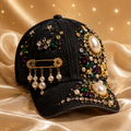 A black baseball cap heavily embellished with gold-tone jewelry, faux pearls, and colorful rhinestones, resting on a draped champagne satin fabric background with sparkling light effects.