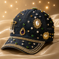A dark navy blue denim baseball cap heavily embellished with gold rhinestones, faux pearls, and green gemstones, resting on a draped beige satin fabric with a soft bokeh light background.