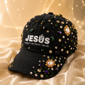 A black baseball cap embellished with gold and multicolor rhinestones and pearl-like beads, featuring the text 'JESUS THE WAY - THE TRUTH - THE LIFE' on the front. The cap is styled on a draped champagne satin fabric with a soft, glowing bokeh background.